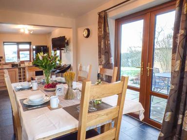 A dining area with wooden chairs and a table set with dishes cups and flowers next to glass doors opening to a patio at Tynddol Bungalow in Llanbadarn-Fynydd near Llanbister