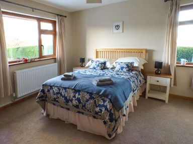 A bedroom with a double bed blue patterned bedspread bedside tables with lamps and windows with curtains at Tynddol Bungalow in Llanbadarn-Fynydd near Llanbister