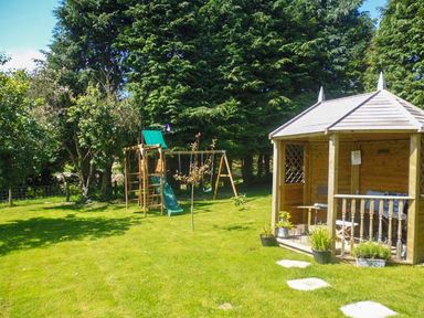 A garden with a wooden gazebo and a children's playset with slide and swings at Tynddol Bungalow in Llanbadarn-Fynydd near Llanbister