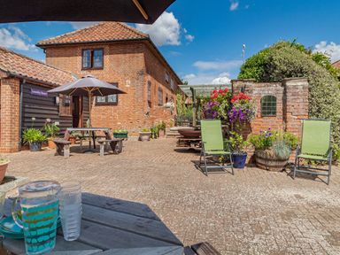 An outdoor patio area with brick buildings green chairs a picnic table with an umbrella and various potted plants at Punch Cottage in Little Glemham near Wickham Market