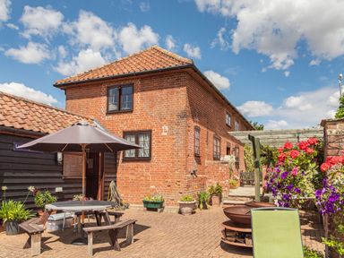 An outdoor patio area with a wooden picnic table and umbrella next to a brick house with multiple windows at Punch Cottage in Little Glemham near Wickham Market