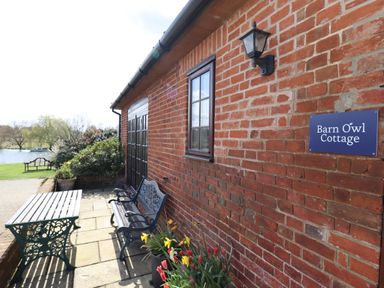An outdoor patio area with a bench and table next to a brick wall with a sign that reads Barn Owl Cottage with flowers and a lawn by water at Barn Owl Cottage in Little Glemham near Wickham Market