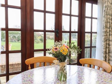 A dining table with a floral tablecloth and a vase of flowers by large wooden framed glass doors with a view of a garden at Barn Owl Cottage in Little Glemham near Wickham Market