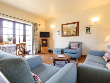A living room with blue sofas a round wooden coffee table a TV on a wooden stand and a dining table with chairs near glass doors at Barn Owl Cottage in Little Glemham near Wickham Market