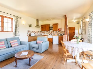A living room with blue sofa and armchair a wooden coffee table kitchen with wooden cabinets and tiled backsplash and a dining table with flowers at Barn Owl Cottage Little Glemham near Wickham Market