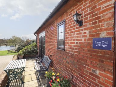 A patio with a bench table and flowers outside a brick cottage by a lake at Barn Owl Cottage in Little Glemham near Wickham Market