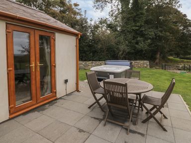 A patio with a round wooden table and five wooden chairs near a hot tub and wooden double doors at Y Beudy Cottage in Clynnog Fawr