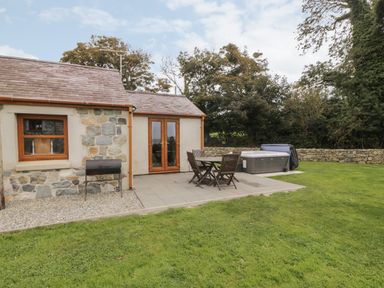 A patio area with wooden table and chairs a barbecue and hot tub outside a stone and plaster cottage at Y Beudy Cottage in Clynnog Fawr