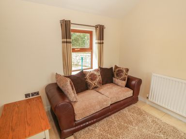 A small sitting area with a sofa cushions window with curtains a wooden side table and a radiator at Y Beudy Cottage in Clynnog Fawr