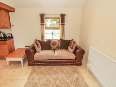 A living room with a brown leather and fabric sofa with cushions a small wooden side table a window with curtains and a radiator at Y Beudy Cottage in Clynnog Fawr