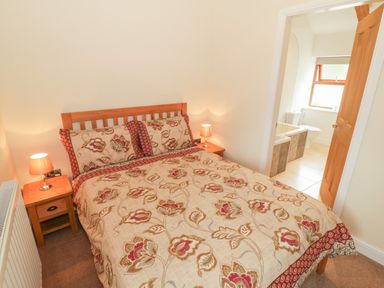 A bedroom with a double bed floral bedding two bedside tables with lamps and a bathroom visible through an open door at Y Beudy Cottage in Clynnog Fawr