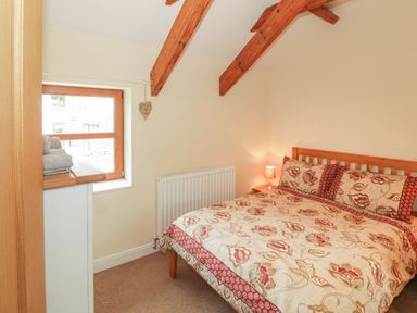 A bedroom with a wooden bed and floral bedding a bedside table with a lamp a window with wooden frame and exposed wooden beams on the ceiling at Y Beudy Cottage in Clynnog Fawr