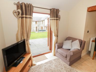 A small living room with a TV on a wooden stand an armchair with two cushions and glass doors with curtains leading to a garden at Y Beudy Cottage in Clynnog Fawr