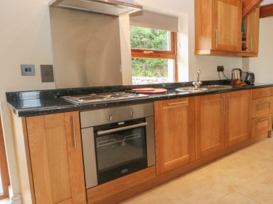 A kitchen with wooden cabinets a black countertop a window oven and stovetop at Y Beudy Cottage in Clynnog Fawr