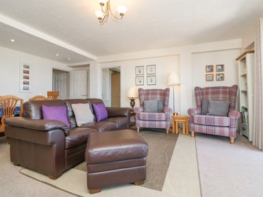A living room with a brown leather sofa and ottoman two plaid armchairs wooden dining chairs and framed pictures on the walls at 37 Ocean Park in Westward Ho