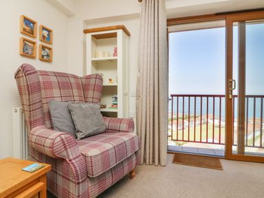 A chair with cushions next to a wooden side table and a white shelving unit by a sliding door with a view of houses and the sea at 37 Ocean Park in Westward Ho