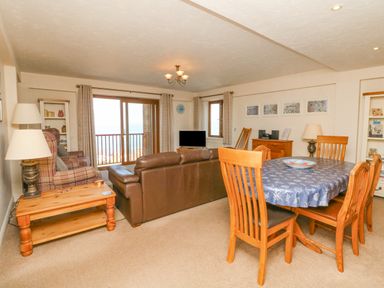 A living room with a dining table covered with a tablecloth a brown leather sofa a plaid armchair a TV and wooden furniture at 37 Ocean Park in Westward Ho