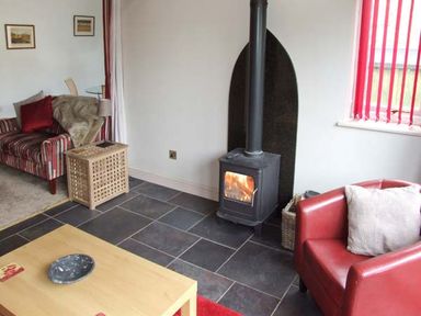 A living room with a wood burning stove a red armchair and a striped sofa at Hour Cottage in Stow-on-the-Wold