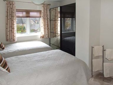 A bedroom with two beds a window with curtains a mirrored wardrobe and a towel rack with towels at Hour Cottage in Stow-on-the-Wold