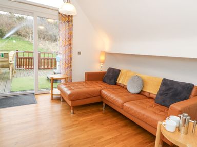 A living room with a brown leather sectional sofa with cushions and a throw next to a sliding door leading to a deck with outdoor furniture at Ty Henri in Tresaith
