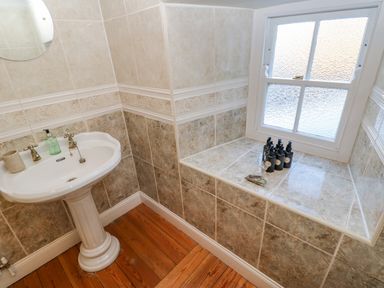 A bathroom with a pedestal sink a round mirror wooden floor and a frosted window with bottles on the windowsill at East Farm House in Humshaugh near Hexham