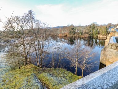 A river with leafless trees along the bank and a stone bridge with a road at East Farm House in Humshaugh near Hexham