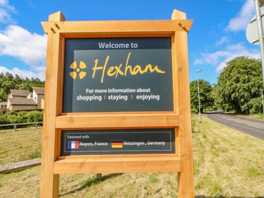 A wooden welcome sign for Hexham located outdoors near a road with trees and houses in the background