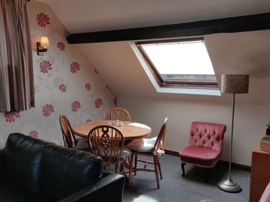 A dining area with a round wooden table and four chairs next to a slanted window and a pink armchair with a floor lamp at The Coach House in North Newton near Bridgwater