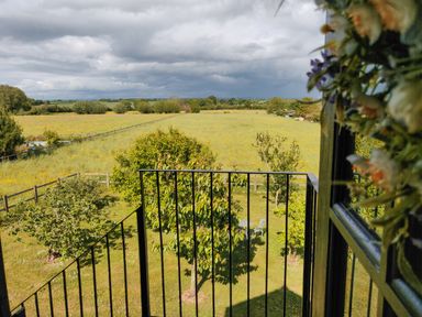 A view from a balcony overlooking a fenced garden with trees and a table and chairs in a rural area