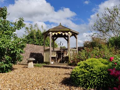 A wooden gazebo with chairs and table in a garden with plants and gravel ground at The Coach House in North Newton near Bridgwater