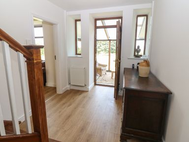 A hallway with wooden stairs on the left a dark wooden cabinet on the right and an open door leading to a sunroom at Y Berth Ddu Farmhouse in Clynnog Fawr