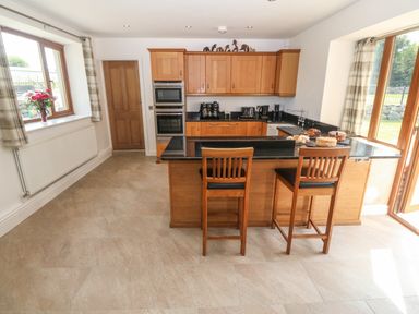 A kitchen with wooden cabinets two bar stools and cakes on the counter at Y Berth Ddu Farmhouse in Clynnog Fawr