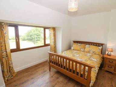 A bedroom with a wooden bed and yellow floral bedding next to a window with matching curtains and a wooden nightstand with a lamp at Y Berth Ddu Farmhouse in Clynnog Fawr