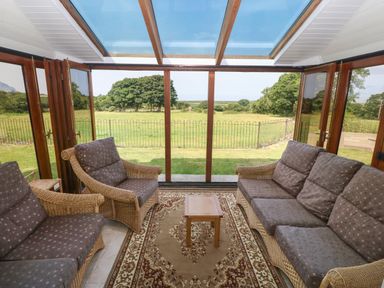 A conservatory with wicker sofas a small wooden table and a patterned rug overlooking a garden at Y Berth Ddu Farmhouse in Clynnog Fawr