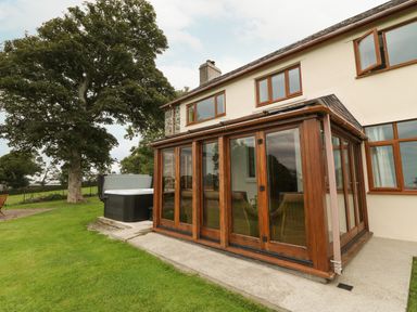 A backyard patio with a glass enclosed wooden conservatory and a hot tub near a large tree at Y Berth Ddu Farmhouse in Clynnog Fawr