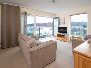 A living room with beige sofas a wooden TV stand and large windows with a view of houses and hills at The Moorings in Deganwy near Llandudno