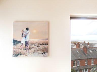 A painting of a couple embracing on a beach at sunset on a wall next to a window showing rooftops of houses and a view of the sea at The Moorings in Deganwy near Llandudno