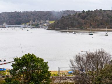 A river with boats anchored near a wooded hillside and a stone castle in the distance at The Moorings in Deganwy near Llandudno