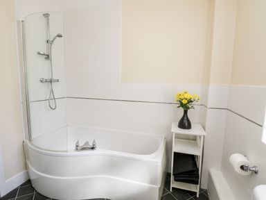 A bathroom with a white bathtub and shower next to a small white shelving unit with a black vase of yellow flowers at Sunnybrae in Whitby