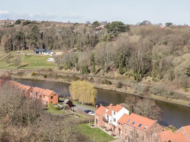 A river flowing beside houses with red roofs and a grassy area with trees behind at Sunnybrae in Whitby