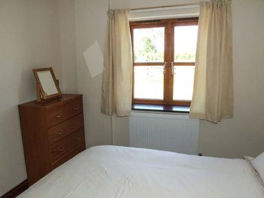 A bedroom with a bed white bedding wooden chest of drawers with a mirror and a window with beige curtains at Dovetail in Westbury-on-Severn