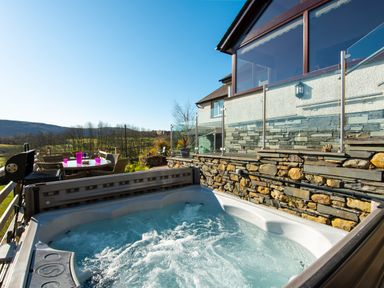 An outdoor hot tub next to a stone wall and glass railing with a patio dining set and barbecue grill in the background at Browside Little Arrow near Coniston