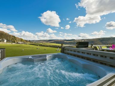 An outdoor hot tub with bubbling water overlooking a green field with sheep and hills in the background at Browside Little Arrow near Coniston