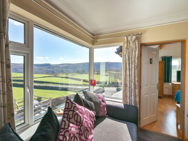 A corner sofa with cushions by large corner windows showing green fields outside and an open door leading to a room with wooden furniture at Browside Little Arrow near Coniston