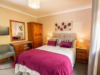 A bedroom with a double bed purple pillows and blanket a wooden wardrobe a dressing table with a mirror a patterned chair and bedside tables with lamps at Browside Little Arrow near Coniston