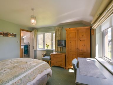 A bedroom with a bed a wooden wardrobe a wooden dresser two chairs a desk and windows with curtains at Browside Little Arrow near Coniston