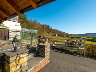 A stone step entrance with outdoor lanterns leading to a patio with outdoor seating and wooden railing overlooking fields and hills at Browside Little Arrow near Coniston