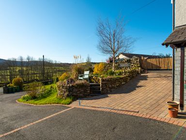 A paved driveway with stone retaining walls and benches next to a lawn with trees and hills in the background at Browside Little Arrow near Coniston