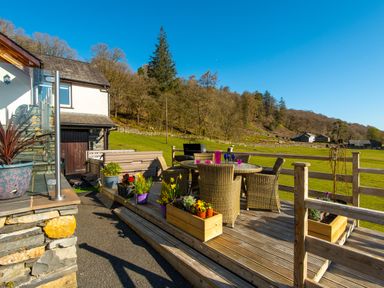 An outdoor wooden deck with a round glass table and six wicker chairs surrounded by flower pots and a hot tub near a house with trees and fields in the background at Browside Little Arrow near Coniston