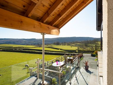 A balcony with glass railing and outdoor furniture overlooking green fields with sheep and hills at Browside Little Arrow near Coniston